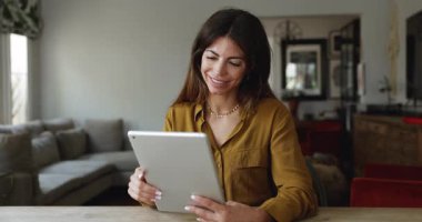 Genç Latin kadın rahat bir oturma odasında oturmuş modern dijital tablet kullanıyor, internette bilgi arıyor, mal arıyor, hizmet satın alıyor, evde tek başına e-alışveriş yapmaktan tatmin oluyor.