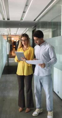Çok ırklı iş arkadaşları iş meselelerini tartışıyor, tablete bakıyor ve işaret ediyor, konuşuyor, ofis koridorunda duruyor. Proje yöneticisi iş arkadaşından tavsiye istiyor. Dikey çekim