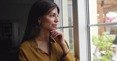 Düşünceli bir ifadeyle dışarıyı seyreden düşünceli Latin bir kadın, bitkilerle ve ağaçlarla dolu bir bahçeyi inceliyor, geleceği düşünüyor, modern evdeki pencerenin yanında tek başına hayal kuruyor.