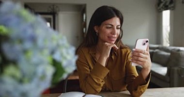 Çekici İspanyol kadın masadaki rahat bir odada oturmuş sevimli çiçeklerle süslenmiş video izliyor, bilgilendirme alıyor, gülümsüyor ve başvuru sırasında sohbet ediyor.