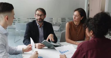 Orta yaşlı takım lideri, veri eğilimlerini göz önünde bulundurarak proje ilerlemesi veya performans ölçümlerini tartışmak üzere modern toplantı odasında oturan çeşitli genç personel üyeleriyle analitik bilgileri paylaşıyor.