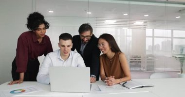 Dört farklı gülümseyen profesyonelden oluşan bir grup ofis masasında dizüstü bilgisayarın etrafında toplandı, ekrana baktılar, dijital iş akışına katıldılar, proje detayları gözden geçirdiler, sonuçları analiz ettiler veya sunumu izlediler.