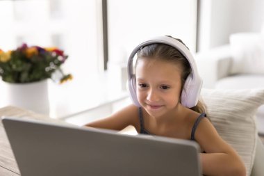 Çocuklar için dijital iletişim. Kablosuz kulaklık takan genç kız, masama otur ve gülümseme eğitimi videosu ile bilgisayar ekranına bak. Oyun oynamakla meşgul olan öğretmen arkadaşınla görüntülü sohbet yap.