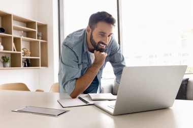 Ev ofisinden öğreniyorum. Mutlu gülümseyen genç erkek öğrenci çalışma masasının yanında duruyor Webinar online eğitim videosu e-öğrenim platformu üzerine sanal sınıf çalışması.