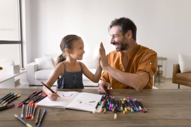 Sanattaki başarıyı kutluyoruz. Heyecanlı küçük kız çocuğu gülümseyen orta yaşlı babaya beşlik çakar yaratıcı hobisinde ebeveynlerin desteğini hissettiği için mutlu olur çizim defterine renkli bir resim çizer.