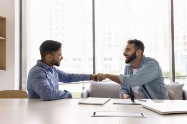 Takım çalışması ve başarı. Farklı etnik kökenlere sahip gülümseyen genç erkek takım arkadaşları iş yerinde yumruk tokuşturma başarılı işbirliğini kutluyor karşılıklı saygıyı hissedin takım ruhu