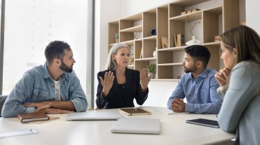 Akıl hocalığı ve işbirliği. Deneyimli kıdemli bayan takım lideri patron iş koçu eğitmeni odaklanmış genç stajyerlerle, yönetim kurulu masasında çeşitli çok kültürlü ofis çalışanlarıyla konuşuyor.