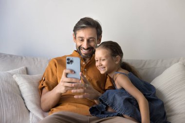 Eğitim ve eğlence için dijital teknoloji. Yetişkin, bekâr bir baba ve küçük bir kız çocuk oturma odasındaki rahat koltukta sarılıyor cep telefonuyla komik aile selfisi çekiyor interaktif oyun oynuyor.