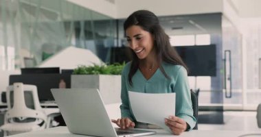 Olumlu genç Latin CEO 'su iş yerindeki kağıt finansal raporunu analiz ediyor, bilgisayardaki muhasebe servisini kurumsal internet iletişimi için kullanıyor, dizüstü bilgisayarda yazıyor, gülümsüyor, yüzünü çeviriyor.