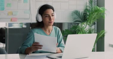 Pozitif genç İspanyol ürün yöneticisi kulaklıklı kadın dizüstü bilgisayardaki şirket toplantısında konuşuyor, video konferans görüşmesinde konuşuyor, kağıt pazarlama raporuna danışıyor.