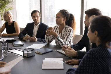 İş görüşmesi. Genç Siyahi kadın, yönetim kurulu masasında çeşitli çoklu etnik takım arkadaşlarıyla brifing hakkında fikir beyan ederek şirketin beyin fırtınasında büyümesini finanse etmek için yatırım payı planı sunuyor.