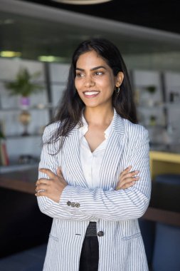 Şık çizgili ceket giymiş kendine güvenen genç Hintli kadın modern ofiste kollarını kavuşturarak duruyor, özgüven, liderlik ve iş yerinde başarı, dikey portre