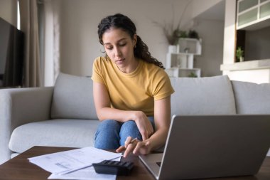 Yurt dışından mali işler yapan bir öğrenci. Meşgul genç kız kanepede oturup kağıt dökümanlarını analiz ediyor defter hesap makinesi kullanarak harcama planlama bütçelerini takip ediyor faturaları online olarak ödemek için