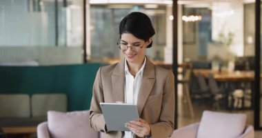 Ciddi anlamda başarılı genç iş kadını, gözlüklü, tablet yazan, ofis lobisinde bir aletle duran, finansal online başvuru kullanan, iş işleri için web servisini kullanan, mutlu olan, gülümseyen