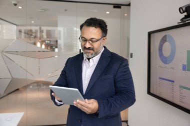 Yetişkin Hintli profesyonel işadamı sunumu gözden geçiriyor, toplantıdan önce slaytları veya notları kontrol ediyor, tablet stantları kullanarak büyük dijital ekran görüntüleme grafikleri kullanıyor. Veri analizi, iş akışı, teknoloji