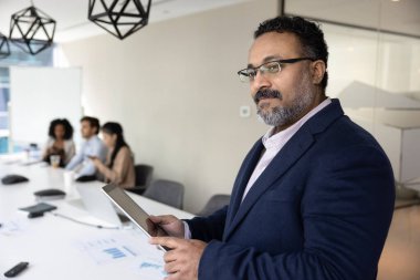 Düşünceli orta yaşlı Hintli işadamı dijital tabletini kullanarak toplantı odasında duruyor, verileri gözden geçiriyor, gelecek toplantı için sunum hazırlıyor veya hissedarlarla görüşmeler yapıyor. Modern teknoloji