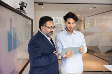 Modern yönetim kurulu toplantısında tablet üzerinde gösterilen performans ölçümlerini analiz eden iki odaklı işadamı, analitik verilerle dijital ekranın yanında duruyorlar. İş dünyasında yenilikçi teknoloji, analitik