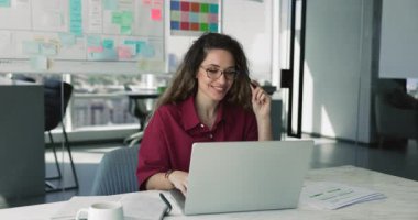 Ofiste tek başına çalışan, iş yerinde dizüstü bilgisayarda yazan, görev çözümü üzerine düşünen, çevrimiçi proje meydan okuyan, gülümseyen, çeneye dokunan düşünceli genç ürün yöneticisi kadın.