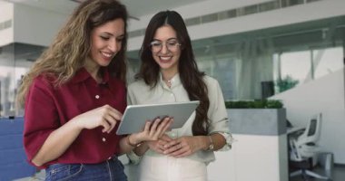 Neşeli genç iş arkadaşları iş molasında tablet kullanıyorlar, ofis yerinde duruyorlar, internete, sosyal medyaya, çevrimiçi mağaza sitesine bakıyorlar, iş ürünlerini tartışıyorlar.