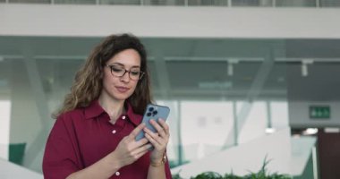 Cep telefonuyla yazan gözlüklü genç ofis çalışanı, ofiste tek başına duran, sosyal medyayı kaydıran, interneti tarayan, online iletişim için aygıt kullanan.