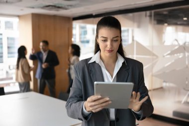 Resmi giyimde ciddi bir kadın ofiste tablet kullanıyor, arka planda meslektaşları var. Profesyonel kadın gözden geçirme projesi güncellemeleri, çevrimiçi sunum hazırlama, birlikte çalışma alanında duran çevrimiçi görev üzerinde çalışma