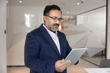 Orta yaşlı Hintli analist, resmi takım elbiseli işadamı modern çalışma ofisinde duruyor, verileri analiz ediyor, e-posta atıyor, sunum hazırlıyor, dijital belgeyi veya tablet hakkındaki mali raporu inceliyor