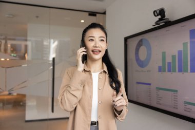 Modern ofiste telefon görüşmelerine katılan Asyalı profesyonel kadın, ekranın yanında duruyor, çeşitli grafikleri gösteriyor, gülümsüyor, iş performansını tartışıyor, gelir artışını tartışıyor veya veri analizi yapıyor.