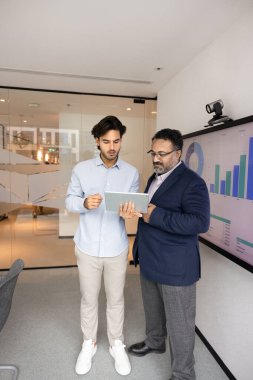 İki çok ırklı, genç ve olgun profesyonel toplantı odasında büyük dijital ekran grafiklerinin yanında duruyor, mali verileri veya tartışma ve takım çalışmasında kullanılan tablet üzerindeki analizleri gözden geçiriyorlar.