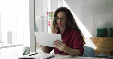 İş için tablet üzerinde online hizmet kullanan gözlüklü, iş yerindeki dijital cihazlarda interneti karıştıran, sosyal medyayı kaydıran olumlu, genç ve güzel bir kadın.