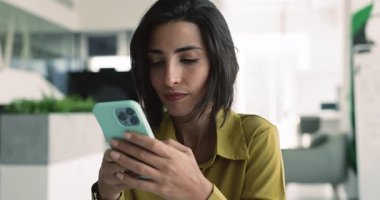 Ciddi genç İspanyol iş kadını ofiste akıllı telefon kullanıyor kurumsal iletişim için, cep telefonuyla yazı yazıyor, iyi haberler okuyor, mutlu oluyor, neşeli oluyor, gülüyor.