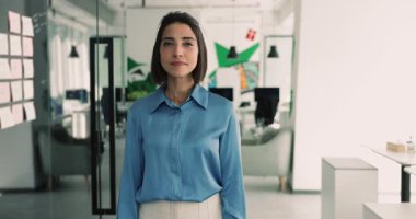 Pozitif, genç ve güzel İspanyol iş kadını mutlu oluyor, gülümsüyor, kameraya bakıyor, güven jesti olarak kollarını göğsüne kavuşturuyor, modern ofiste kargaşa tahtasında poz veriyor. Kafasından vurulmuş.