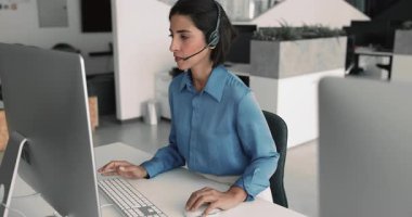 Genç Latin müşteri destek servisi çalışanı video görüşmesinde konuşuyor, ofiste mikrofon ve masaüstü monitörüyle kablosuz kulaklık kullanıyor, online danışmanlık yapıyor.