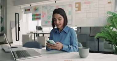 Çevrimiçi iş iletişimi için dijital cihazlar kullanan, cep telefonu ve laptop kullanan, kağıt satış raporlarını kontrol eden mutlu genç Latin iş kadını.