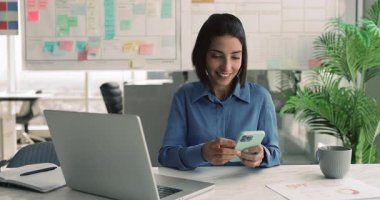 İş yerinde akıllı telefondan mesaj atan, laptopta yazan, iş yerinde konuşmak için farklı aletler kullanan mutlu, genç İspanyol iş kadını.