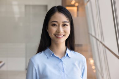 Asyalı bir iş kadınının, sıradan mavi gömlekli, neşeli ve iyimser bir ifadeyle kameraya bakarken çekilmiş resmi. Şirket çalışanlarının ofis portresi, yetenekli kadın çalışanlar.