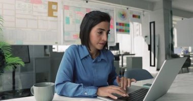 Ciddi odaklı genç İspanyol iş kadını ofiste dizüstü bilgisayarla yazı yazıyor, notlar yazıyor, mutlu oluyor, gülümsüyor, proje geliştirme üzerinde çalışıyor.