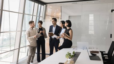 Beş ciddi odaklı takım arkadaşı, ortak proje tartışması için gökdelen ofisinde toplandı. Avrupalı ekip üyesi konuyu açıklıyor, brifing sırasında yeni fikirleri ve yaratıcı çözümü paylaşıyor.