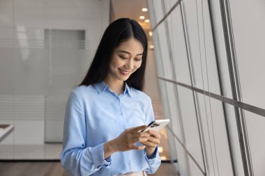 Modern ofiste akıllı telefon kullanan gülümseyen Asyalı iş kadını, yazışmaları kontrol et, e-posta at, iş mesajlarına cevap ver, yeni cep telefonu uygulaması öğren.