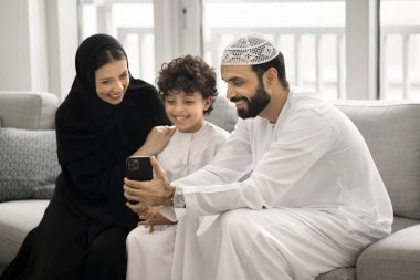 Modern teknoloji ve aile anları. Geleneksel Müslüman giysileri içinde küçük bir oğlu olan mutlu, evli, Arap bir çift oturma odasındaki kanepede otururken cep telefonu kullanmaktan zevk alıyor.