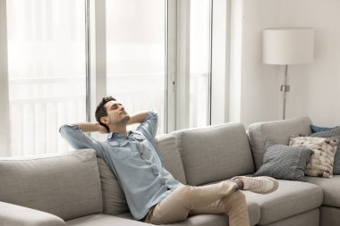 Hayatla barışık. Kaygısız, sakin genç adam rahat kanepeye uzanmış, gözleri kapalı, huzurlu bir uyku molası veriyor modern oturma odasında dinleniyor, kendi evinde huzur içinde sessiz meditasyon müziğini dinliyor.