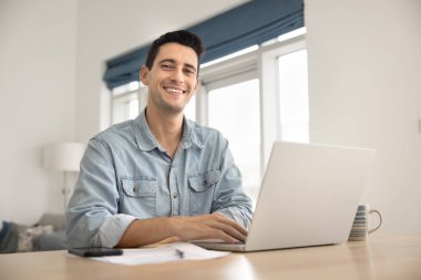 Mesafeli çalış ya da çalış. Mutlu portre mutlu genç adam serbest çalışan uzak öğrenci ev ofisinin masasında oturuyor evrak işleri için dijital teknolojiyi kullanıyor kendine güvenen bir gülümsemeyle kameraya bak.