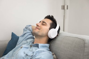 Müzikle rahatlamak. Kablosuz dijital kulaklıklı mutlu bir adam yumuşak kanepeye uzanmış en sevdiği melodiyi dinliyor ve huzur içinde evde tek başına vakit geçiriyor.