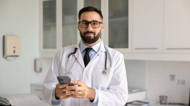 Laboratuvar önlüklü ve gözlüklü genç İspanyol doktor kameraya bakıyor, hastane ofisinde afiş için poz veriyor, cep telefonu kullanıyor.