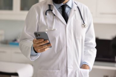 Beyaz önlüklü genç bir doktorun cep telefonuyla yazı yazarken, sağlık hizmetleri iletişimi için başvuru yaparken, hastayla sohbet ederken, uzaktan kumandalı online destek verirken,