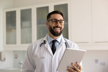 Gözlüklü genç Latin doktor elinde tablet, hastane ofisinde dikiliyor, gülümsüyor, gülüyor, modern dijital teknolojiyi kullanıyor, sağlık hizmetleri için online servis yapıyor.
