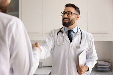 Gözlüklü ve beyaz önlüklü yakışıklı İspanyol doktor hastane ofisindeki iş arkadaşına el sıkışıyor. Saygı gösteriyor, başarılı iş birliğine şükran duyuyor ve el sıkışıyor.