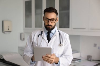 Tıbbi muayene odasındaki tablete yazı yazan, hastalarla sohbetlerde online danışmanlık desteği veren, elektronik muayene raporlarını gözden geçiren, şık gözlüklü ciddi meşgul Latin doktor.