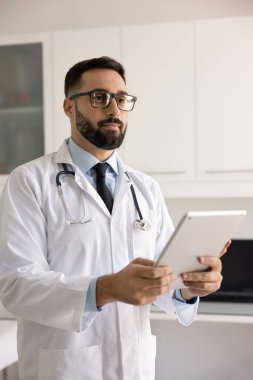 Gözlüklü ve beyaz önlüklü yakışıklı bir doktor, sağlık hizmetleri için tablet bilgisayar kullanıyor, klinikte elinde aletle dikiliyor, başka yere bakıyor, dijital cihaz tutuyor.