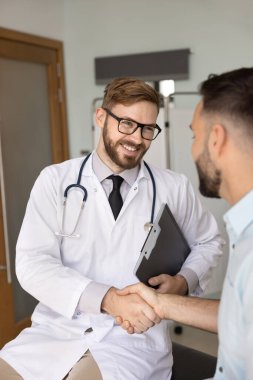 Gözlüklü ve beyaz önlüklü yakışıklı pratisyen, sağlıklı bir hastaya el sıkışarak iyileşmesini kutluyor, başarılı bir terapiden sonra iyi sonuçlar alıyor. Dikey çekim