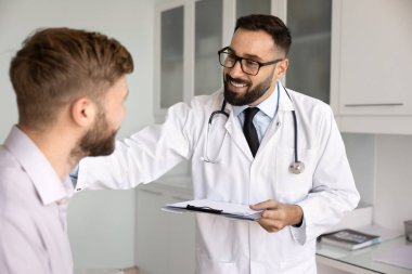 Mutlu İspanyol tıp doktoru genç erkek hastaya iyi haberler veriyor. Omzuna dokunuyor, gülümsüyor, klinik muayene kayıtlarıyla dolu defteri tutuyor, danışmanlık yapıyor.